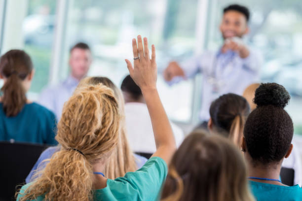 Woman asks question during healthcare seminar Unrecognizable female nurse raises hand to ask a question during medical conference. The conference speaker is pointing at her. lectures in hospital stock pictures, royalty-free photos & images